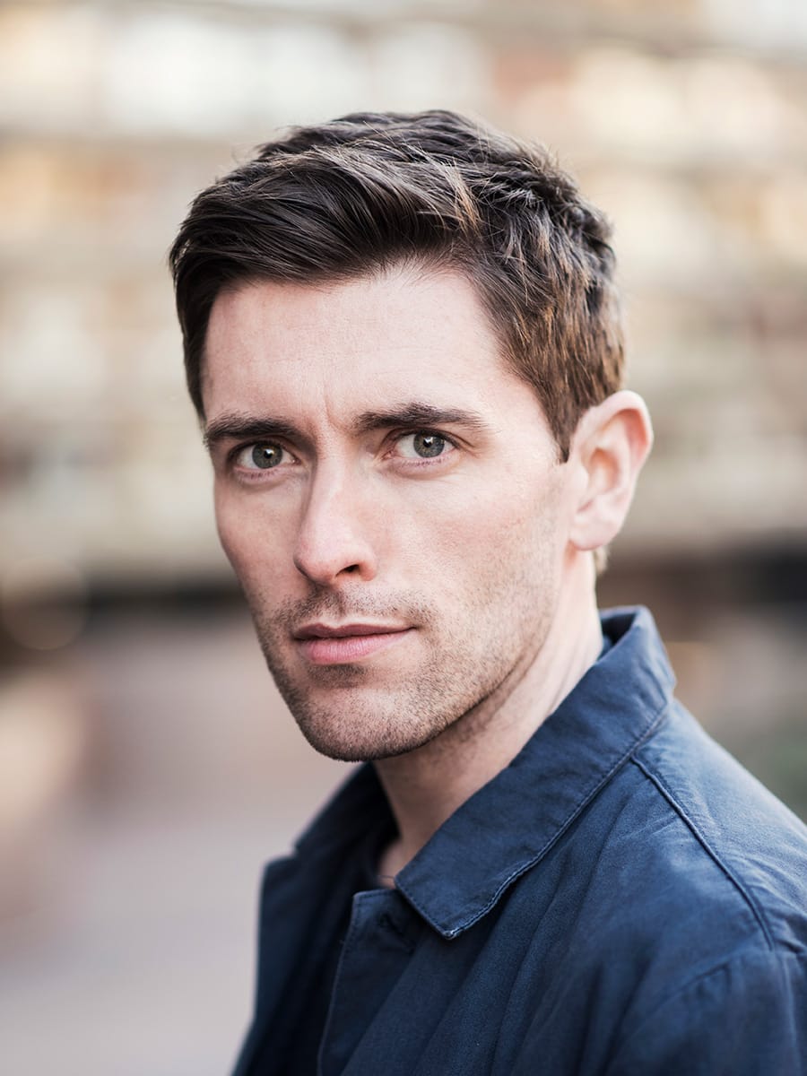 Professional headshot of a man with short brown hair and light eyes, wearing a dark blue collared shirt or jacket. The photo is taken outdoors with a blurred stone or brick background. He has a slight smile and is looking directly at the camera.
