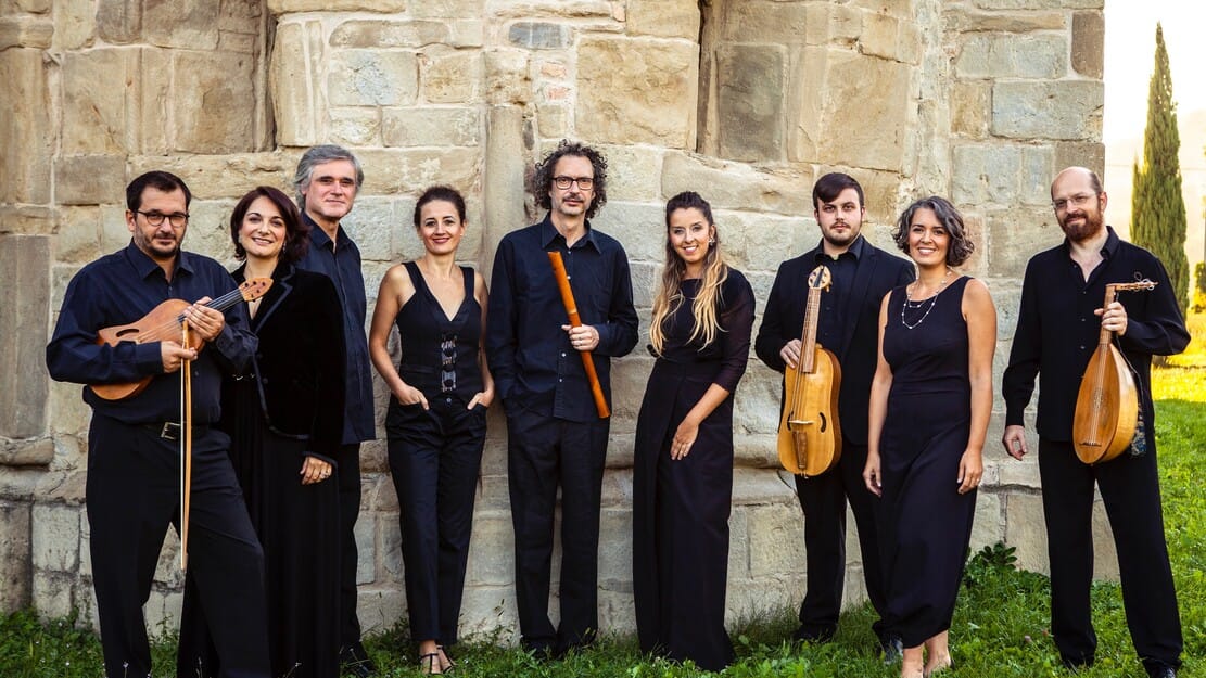 A group of musicians in black attire holding early music instruments, standing outdoors against a stone wall with grass at their feet.