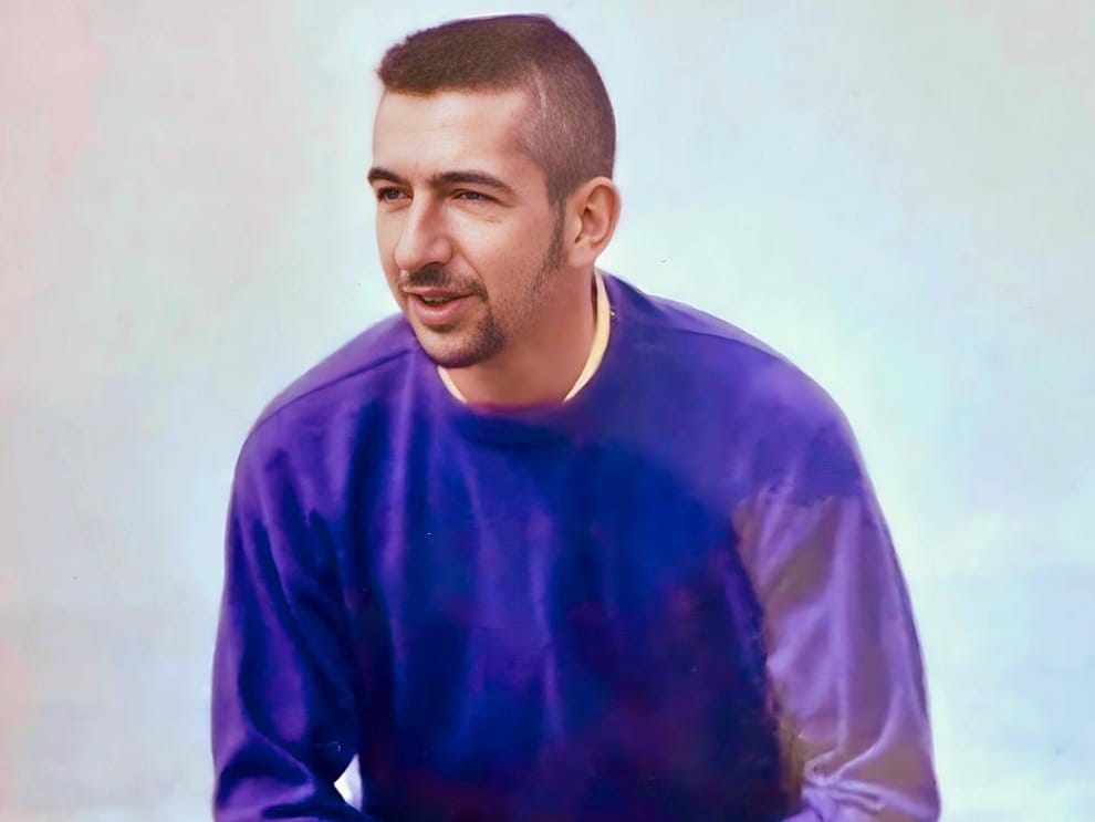 David sits on a bench wearing a purple-blue jumper, rolled jeans, and black boots. He has a sharp fade haircut, neat sideburns, and a brown goatee, looking happy.
