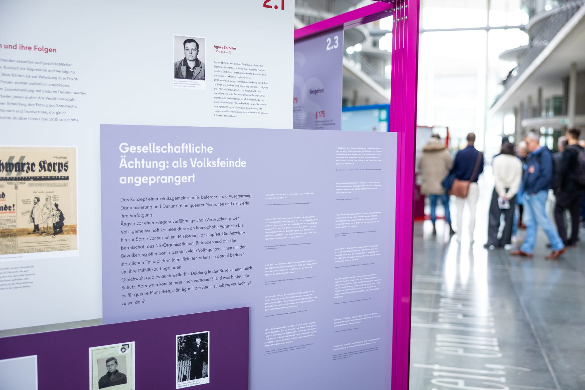 Exhibition panels from the “Endangered Living: Queer People 1933–1945” exhibition, showing historical text, photographs and documents about the persecution of LGBTQ+ people under National Socialism, displayed in a building of the German Parliament with visitors in the background.