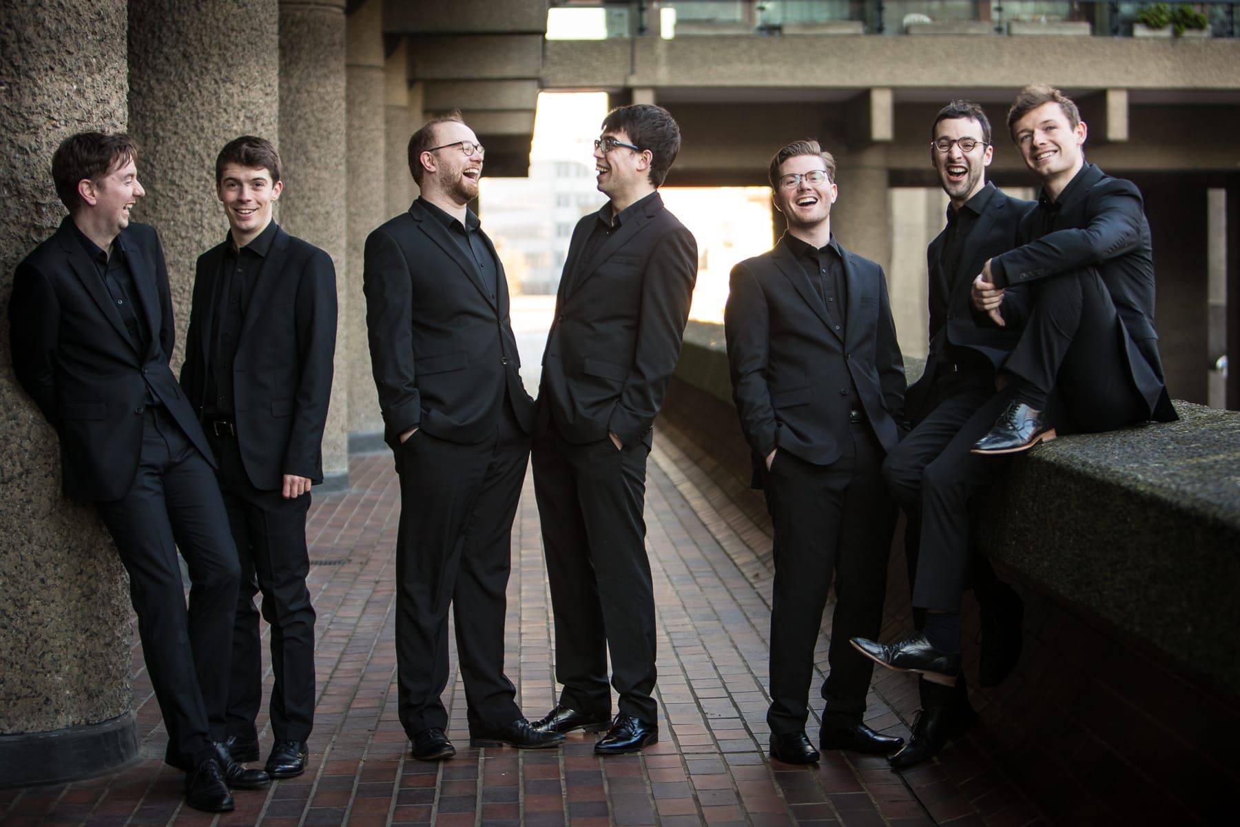 Six handsome men in black suits gathered in a brutalist concrete corridor with brick flooring and concrete pillars, engaged in conversation and smiling.