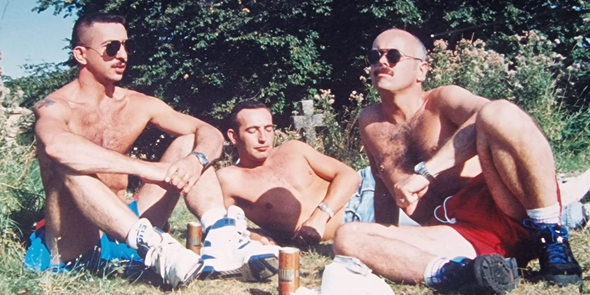 Three gay men relaxing shirtless on grass on a sunny day, wearing sunglasses & shorts, with drinks nearby. The casual photo has a nostalgic film photography quality.