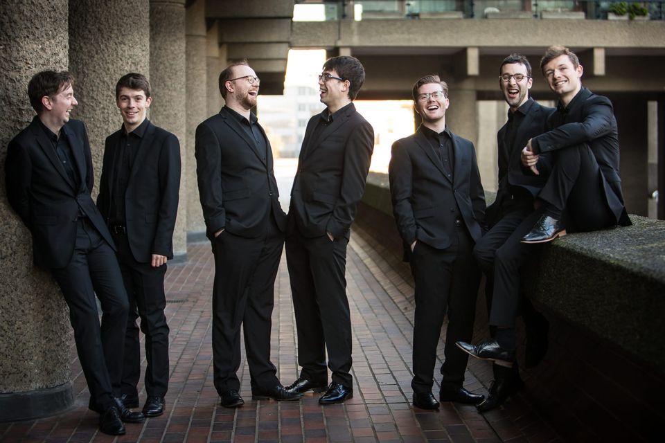 Six handsome men in black suits gathered in a brutalist concrete corridor with brick flooring and concrete pillars, engaged in conversation and smiling.