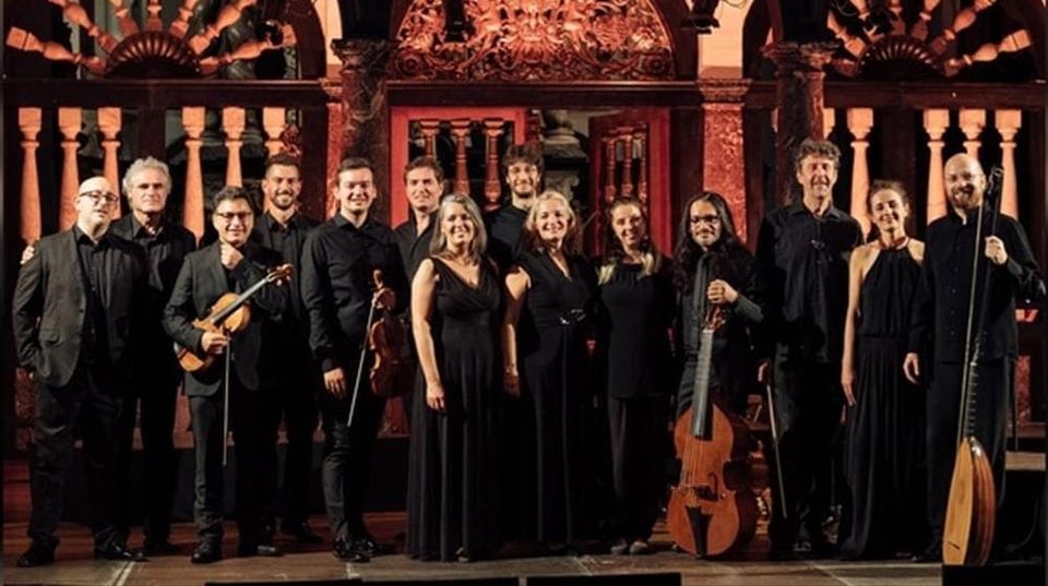 Ensemble of musicians dressed in black, posing with period instruments in front of an ornate, arched backdrop with warm lighting.
