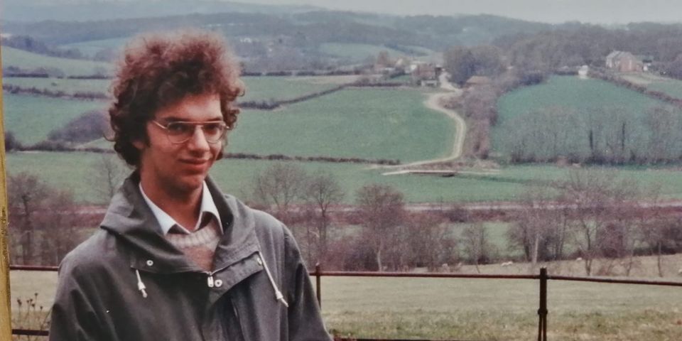 Young man with curly reddish-brown hair and glasses stands in front of a metal gate in the English countryside. He wears a grey jacket over a collared shirt. 