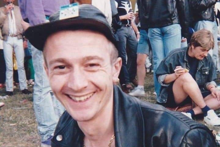 Smiling man in black leather jacket and hat at outdoor gathering, surrounded by people in casual 90s-style clothing at what appears to be a Gay Pride event 1990