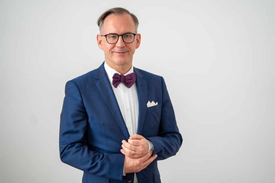 Portrait of Helmut Metzner, Chair of the Federal Magnus Hirschfeld Foundation, wearing a blue suit, white shirt and patterned bow tie, standing against a plain light background.