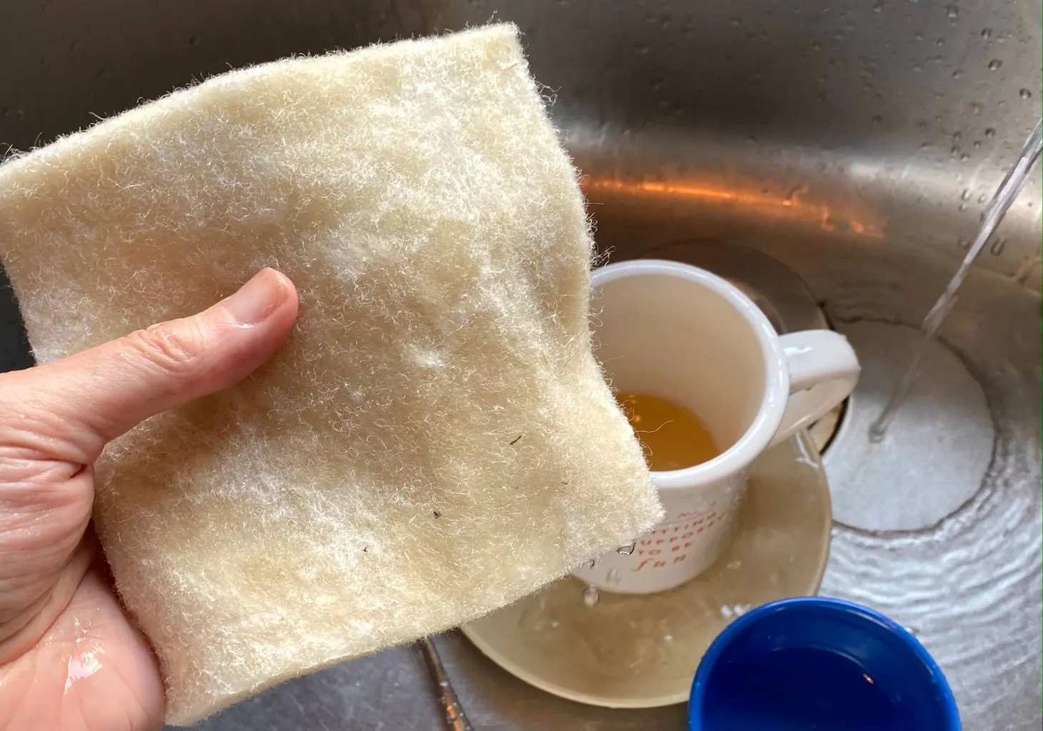 A hand holds a wet square of thick wool felt over a sink of dirty dishes.