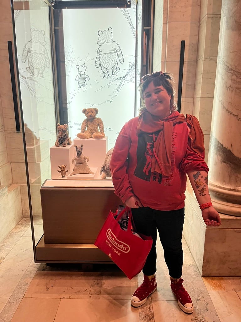 Gay woman standing in orange hoodie and leggings, showing off a tattoo of E.H. Shepard illustrations of Winnie the Pooh characters in front of the original Winnie-the-Pooh stuffed animals behind glass.
