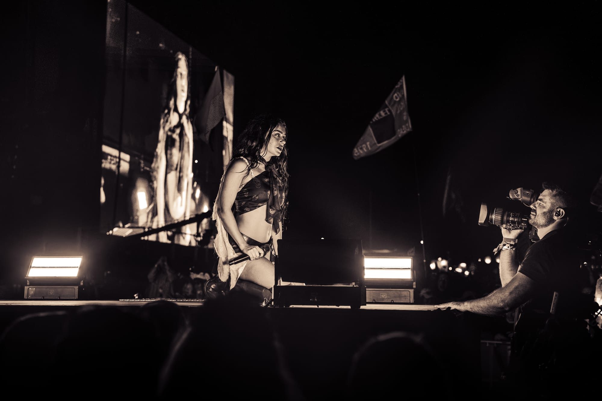 Charli XCX kneeling on stage looking directly into the camera mans lens 