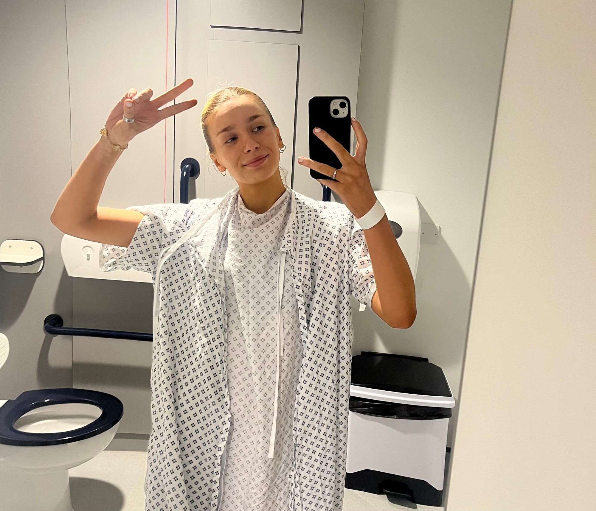 A girl taking a mirror selfie in a hospital bathroom. She's wearing hospital gowns and holding up a peace sign.