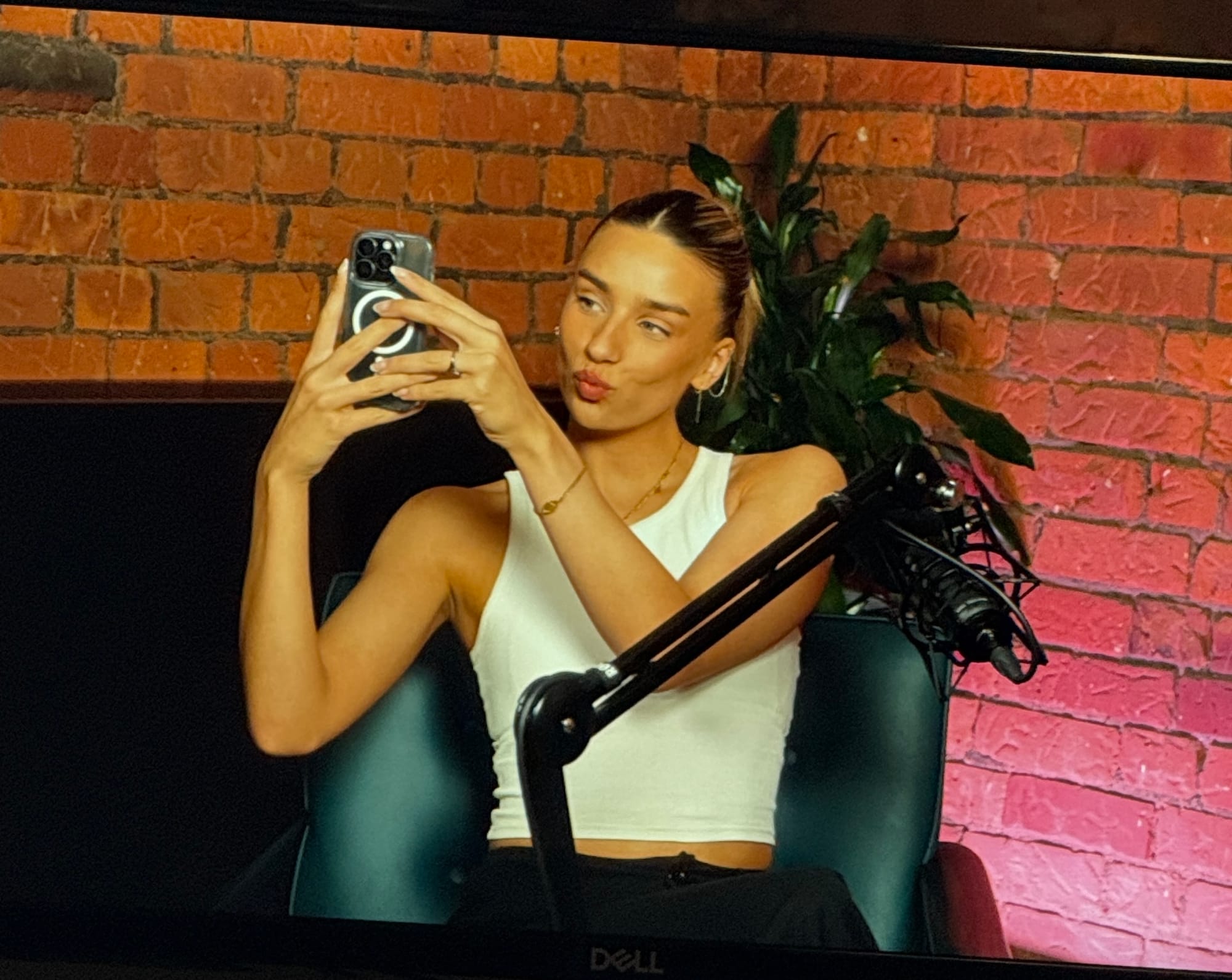 Monitor showing a woman taking a selfie while sitting in a podcast studio with brick walls and professional microphones.