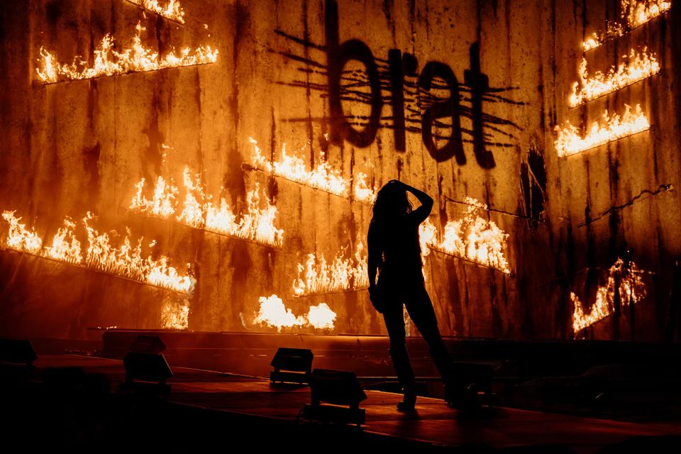 Charli XCX on the Prism Stage at Glastonbury with flames behind her and the 'Brat' logo