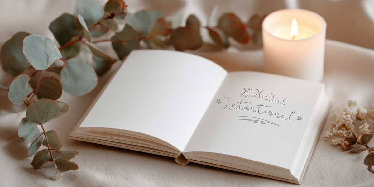 Beige journal open to blank pages reading ‘2026 Word Intentional,’ styled with eucalyptus, a candle, and a small white flower.
