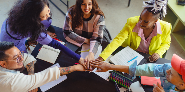 Diverse group of five small business owners around a table and reaching their hands in the centre in a show of support.
