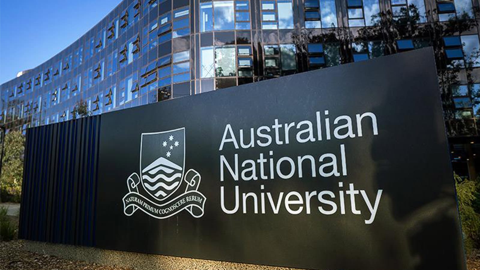 Australian National University sign and crest outside a glass campus building.