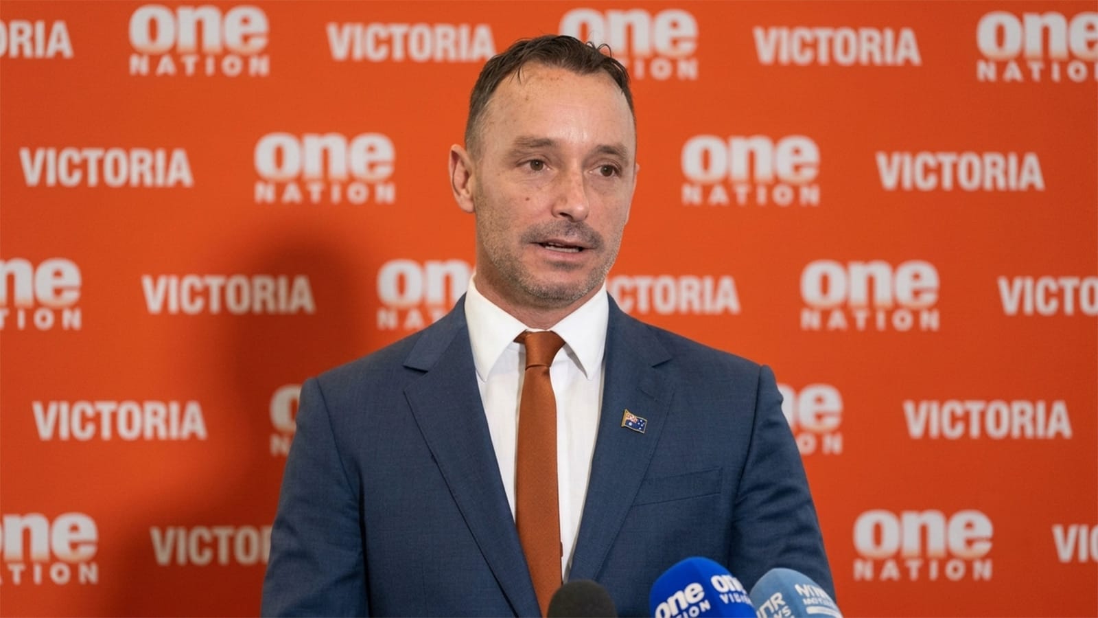 Warren Pickering speaking in front of an orange One Nation backdrop