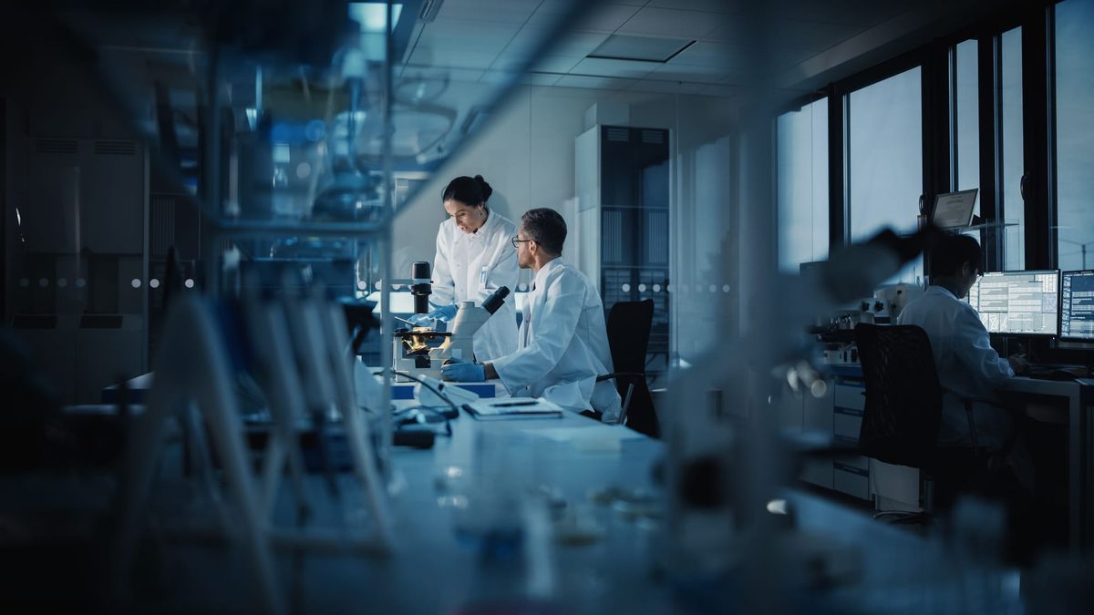 Two scientists talk over a microscope while another works at a computer.