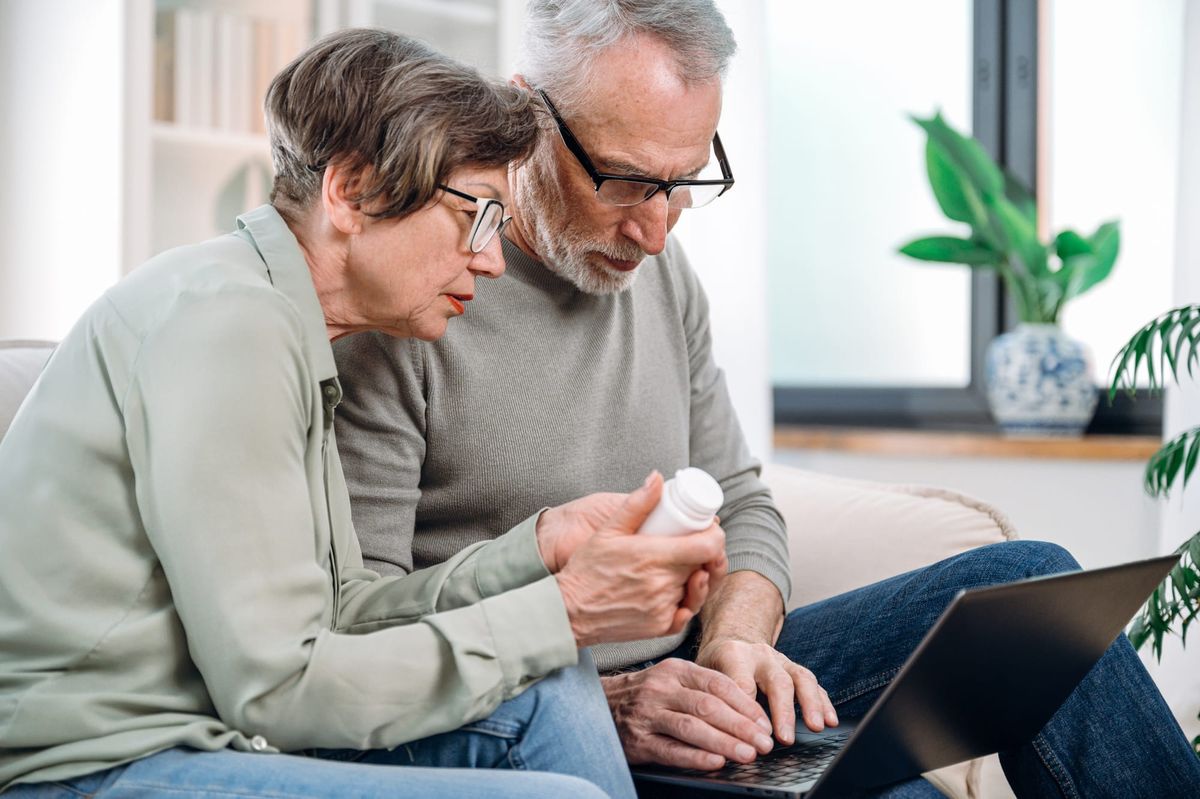 Older couple seated in front of laptop are holding a pill bottle doing research.