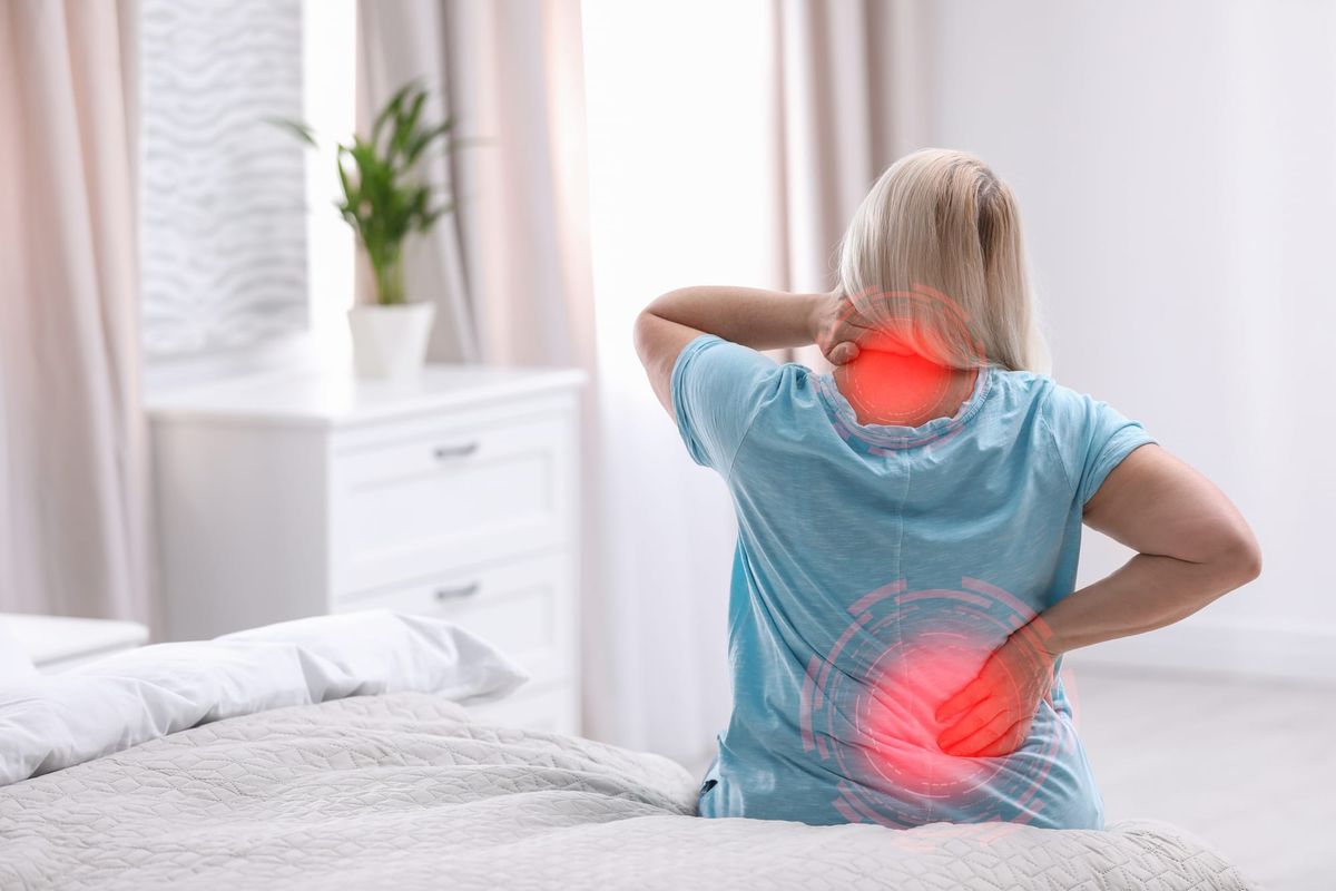 Woman sitting on bed holding her neck and low back which have red circles indicating pain.