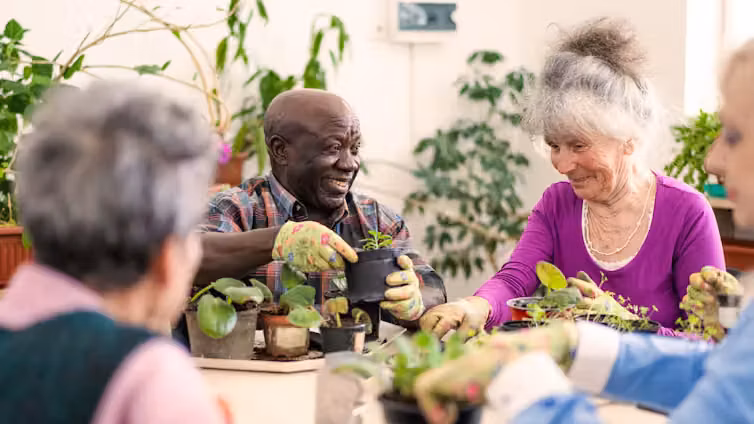 Two smiling older people tend potted plants on a table, with others in the foreground helping out.