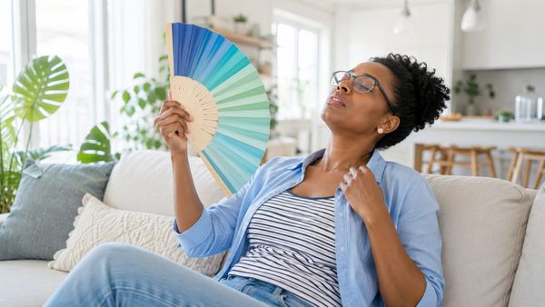 Woman on couch having hot flash fanning herself with a colorful fan.