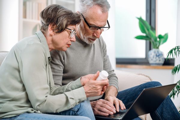 Older couple seated in front of laptop are holding a pill bottle doing research.