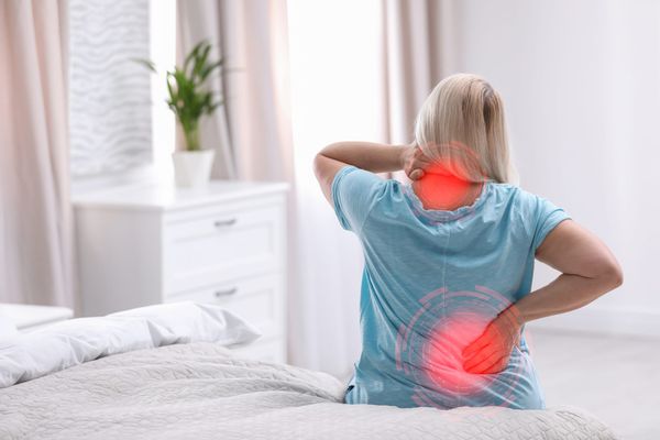 Woman sitting on bed holding her neck and low back which have red circles indicating pain.