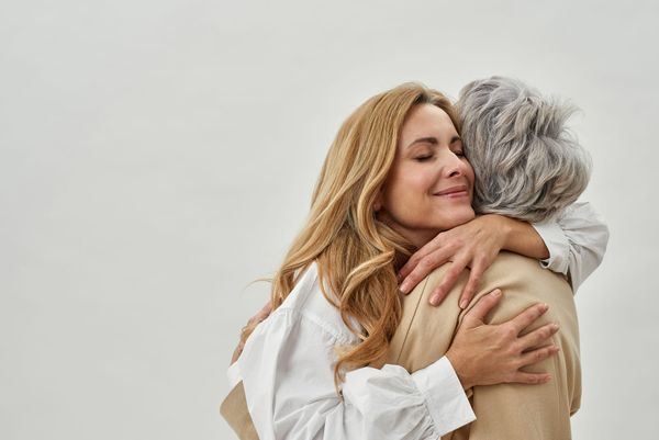 Smiling older woman with eyes closed tightly hugs a gray haired woman. 