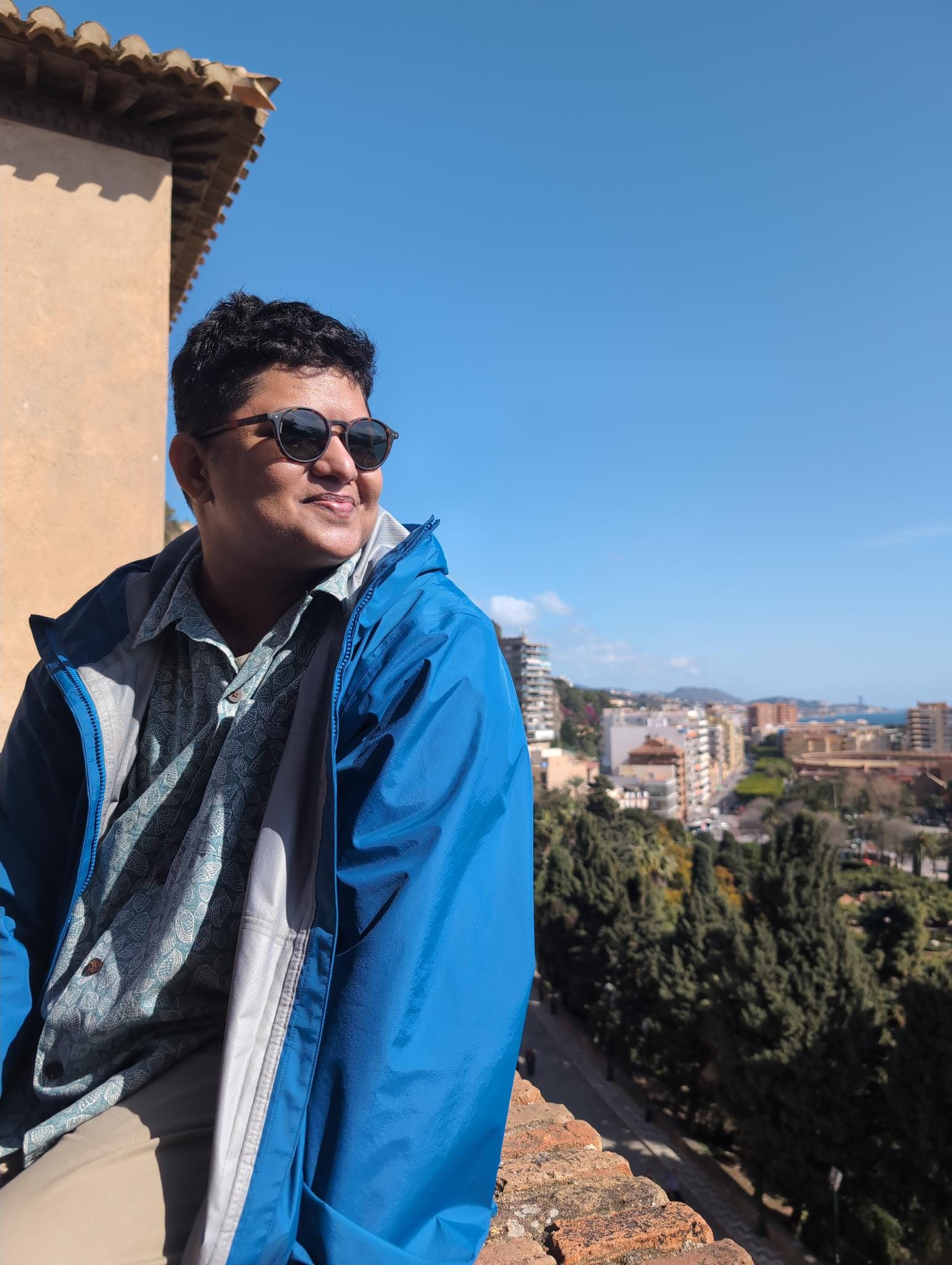 Kadir, a mid-fat brown person wearing sunglasses, blue jacket, light blue shell-print shirt, tan pants, sunglasses sitting on the edge of brick wall overlooking Malaga, Spain with sun on his face