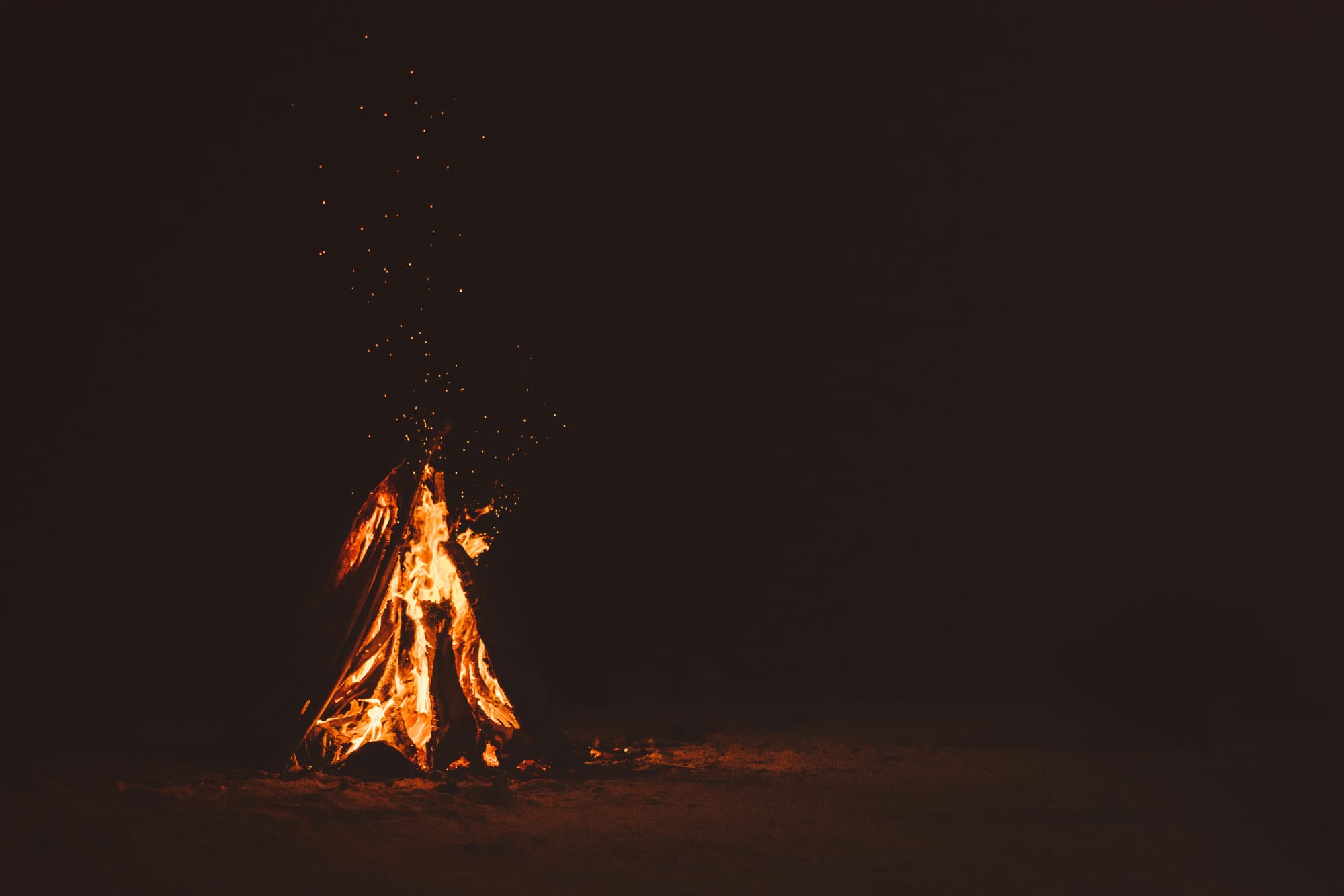 campfire emanating from firewood on sand, dark background, little embers travel towards the sky