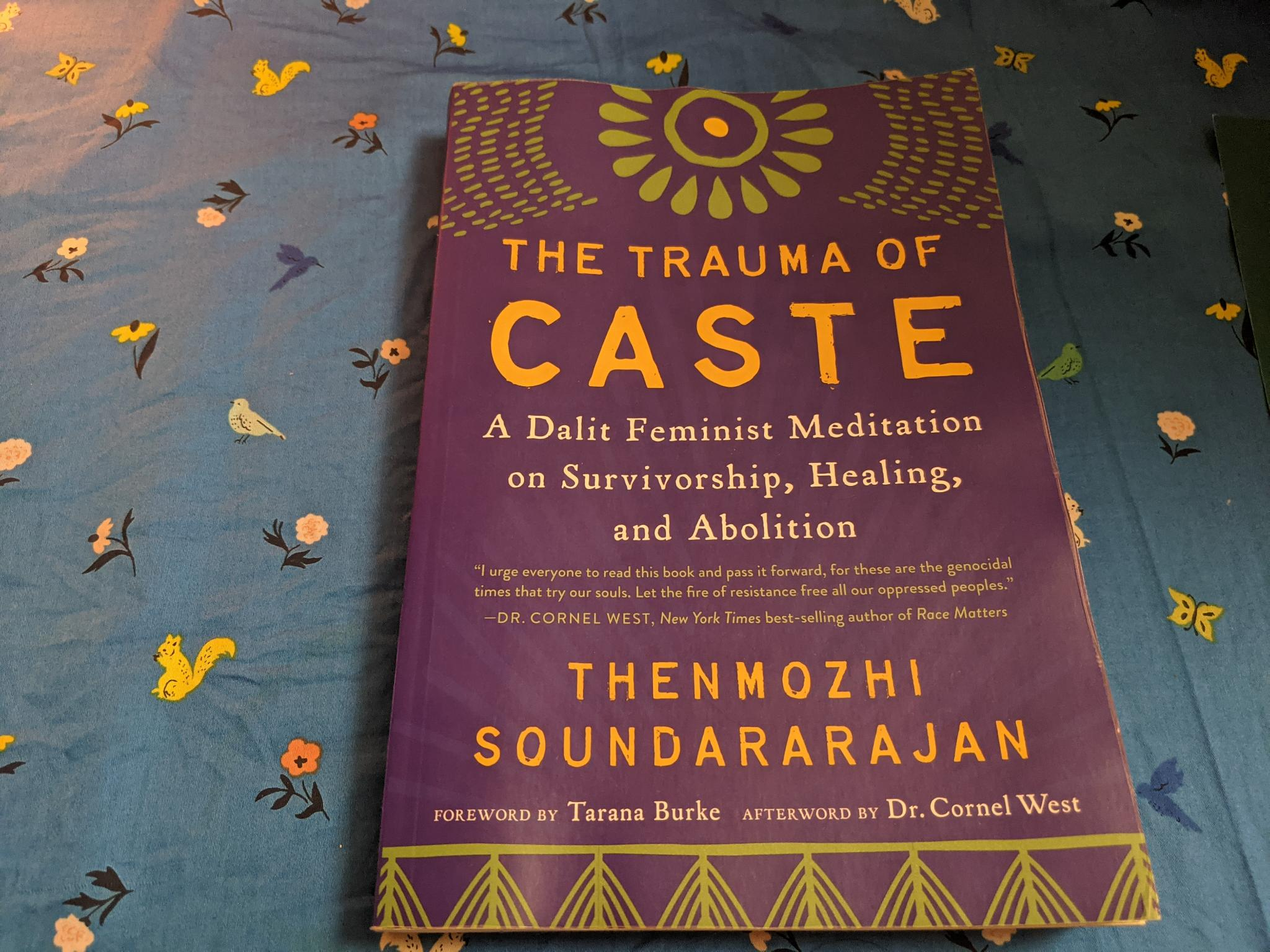 Book cover of "The Trauma of Caste" by Thenmozhi Soundararajan, yellow title text, blue background, green floral print on top,, photographed by Kadir against a blue printed fabric