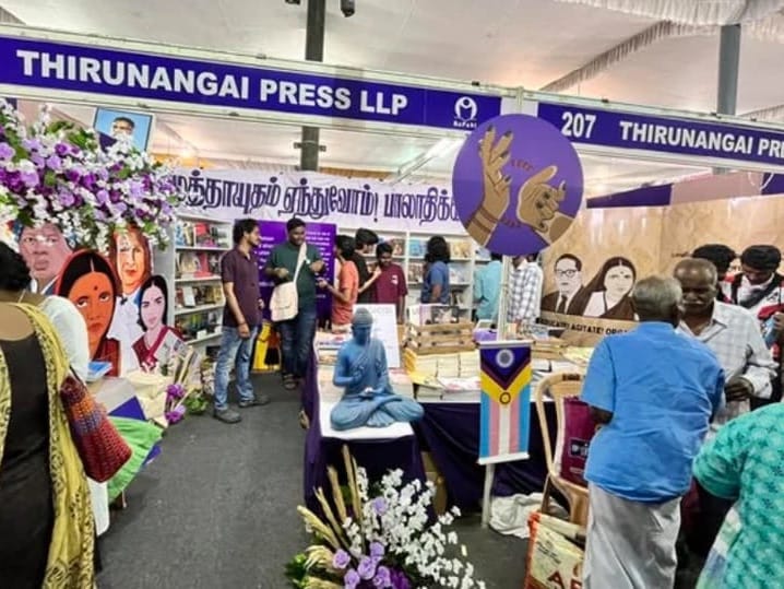 Photo of Thirunangai Press's book stall with blue banner, white lettering on top, at Chennai bookfair 2025, showing people attending and conversing, and cutouts of Dalit icons including Dr. B.R Ambedkar, Ramai Ambedkar, Savithribhai Phule. There is a trans, intersex and genderfluid flag and buddha statue in the middle, and purple and white flower bouquets on the left top and middle bottom of the image. A circular purple background graphic of clapping hands wearing bangles, painted nails is overlayed on the top right-middle of the photo.