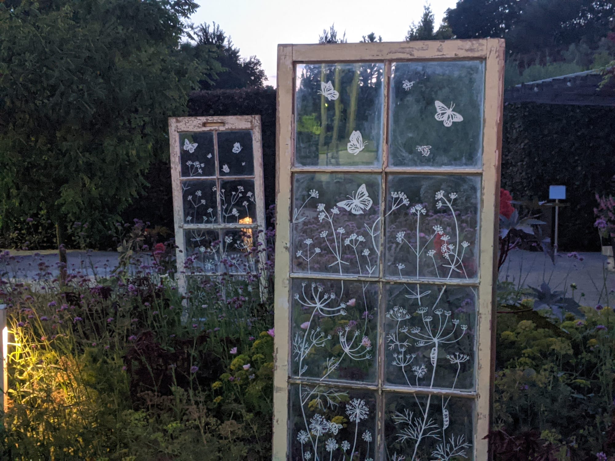 Two beige framed french doors with white flowers and butterflies painted on them, positioned upright amidst a pollinator garden with a variety of colorful flowering plants, with large trees in the background