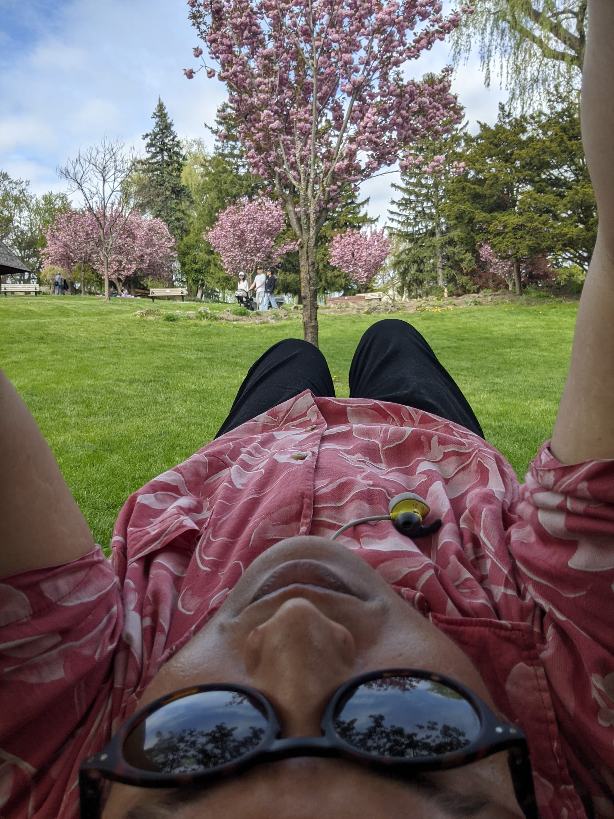 Selfie of Kadir, shot from above, laying on green grass under a flowering tree, wearing red floral print shirt, blue pants and sun glasses.