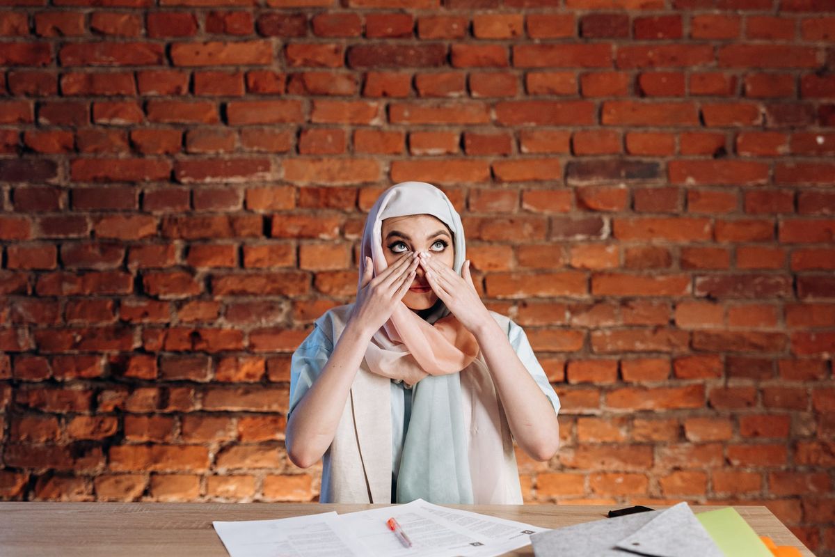 Woman with white hijab wiping tears of their face, setting behind a desk with papers and pens on it, brick wall behind