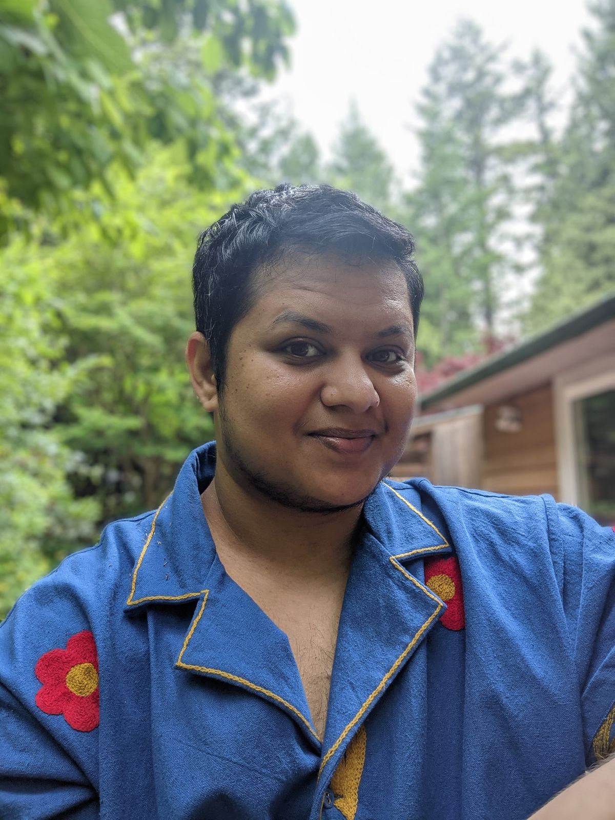 Tan brown skinned nonbinary trans man with black and grey hair, wearing royal blue cotton shirt with yellow and red floral hand embroidery.