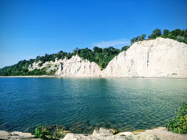 Photo of blue green water overlooking whiteish beige Scarborough bluff hills, greenery and clear blue sky