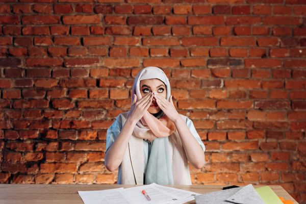 Woman with white hijab wiping tears of their face, setting behind a desk with papers and pens on it, brick wall behind
