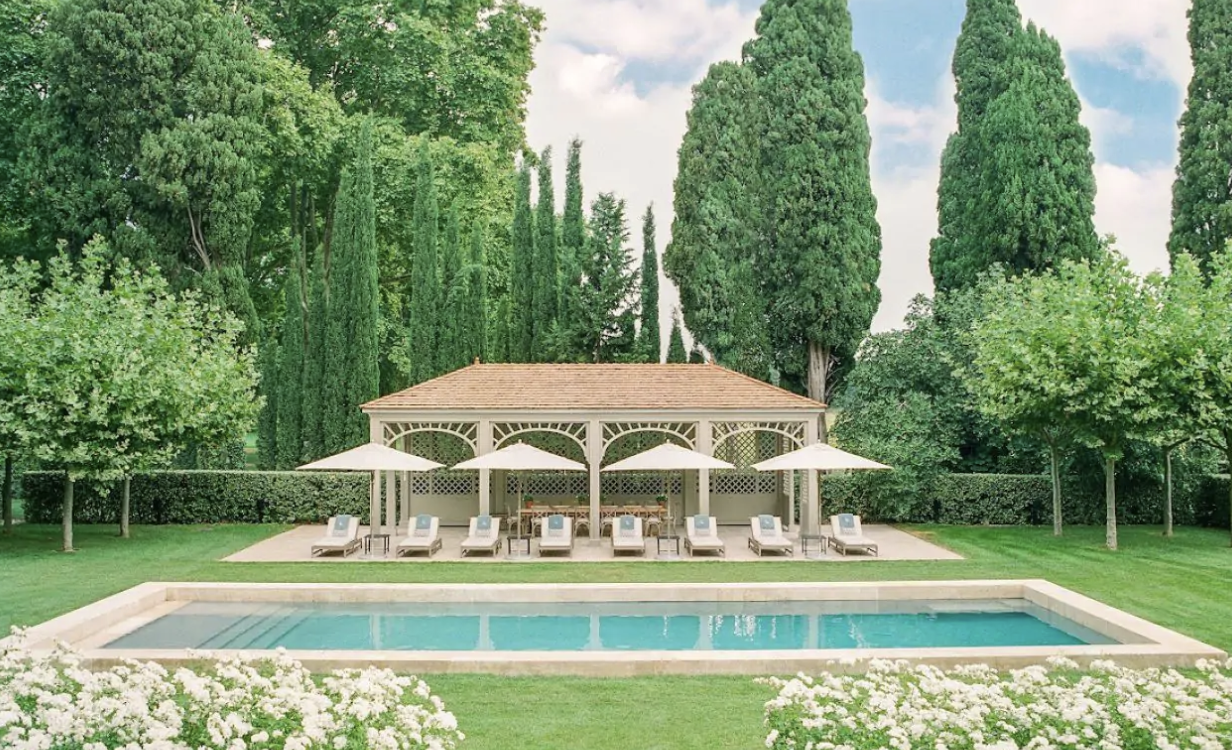  Chateau Avignon’s tranquil swimming pool nestled among lush trees and manicured lawns dotted with sunbeds and umbrellas with a pergola as its focal point.