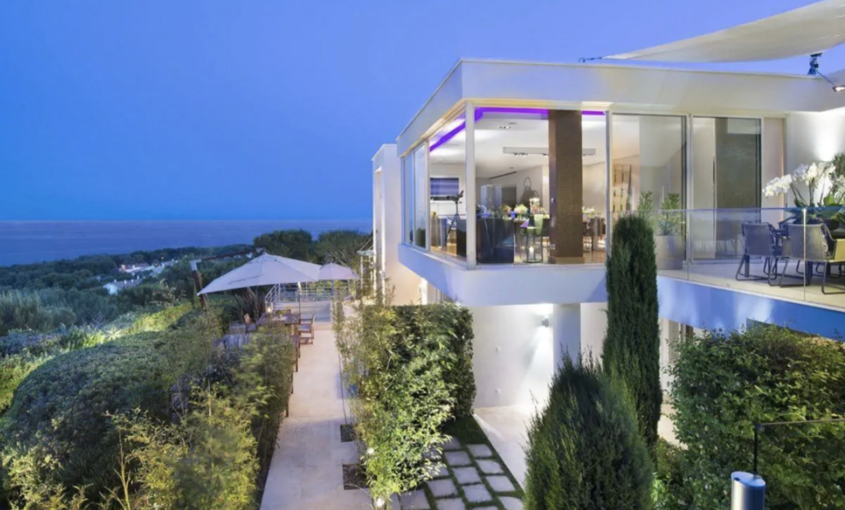 Sleek, modern villa with an expansive terrace and breathtaking ocean views, towering over verdant greenery. 