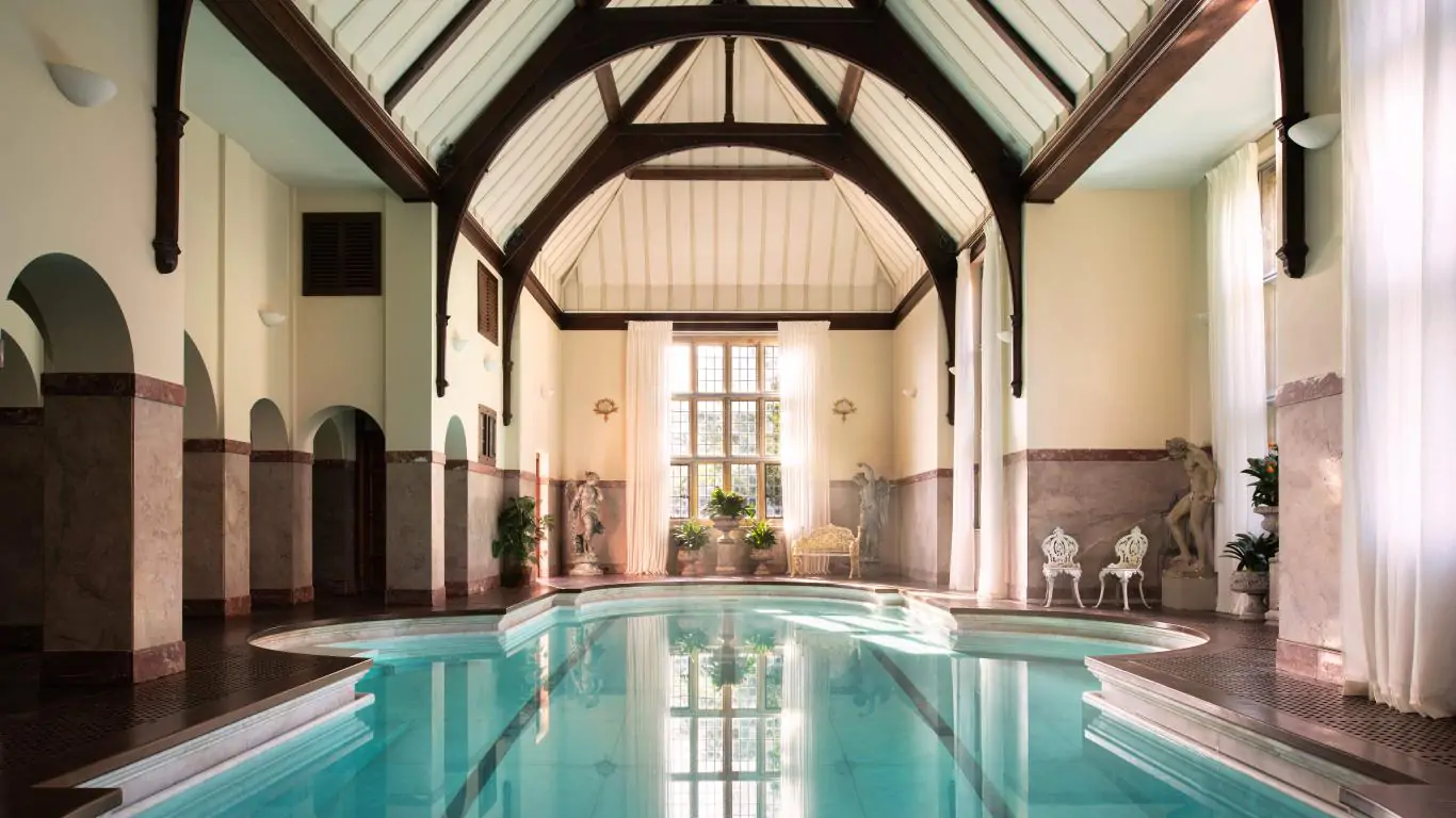 A luxurious indoor pool at Chilham Castle, framed by soaring timber beams, marble accents, and statuesque décor.