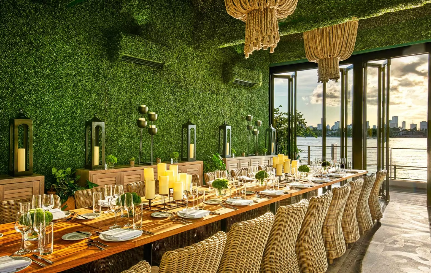 Elegant waterfront dining setup at Baia Beach Club in Miami, featuring a lush green interior, woven chandeliers, and views of the bay.