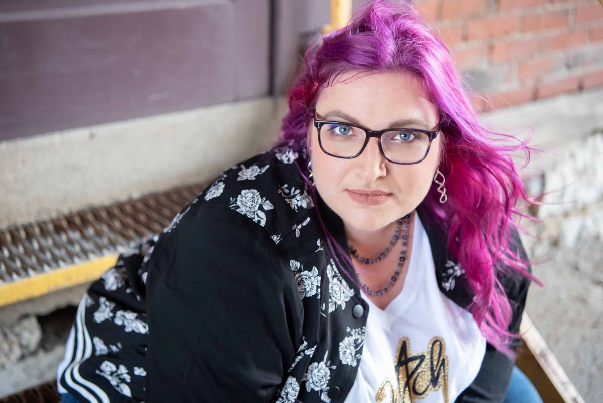 Patty Ryan Lee, with purple hair and glasses, sits in front of a door on metal stairs. 