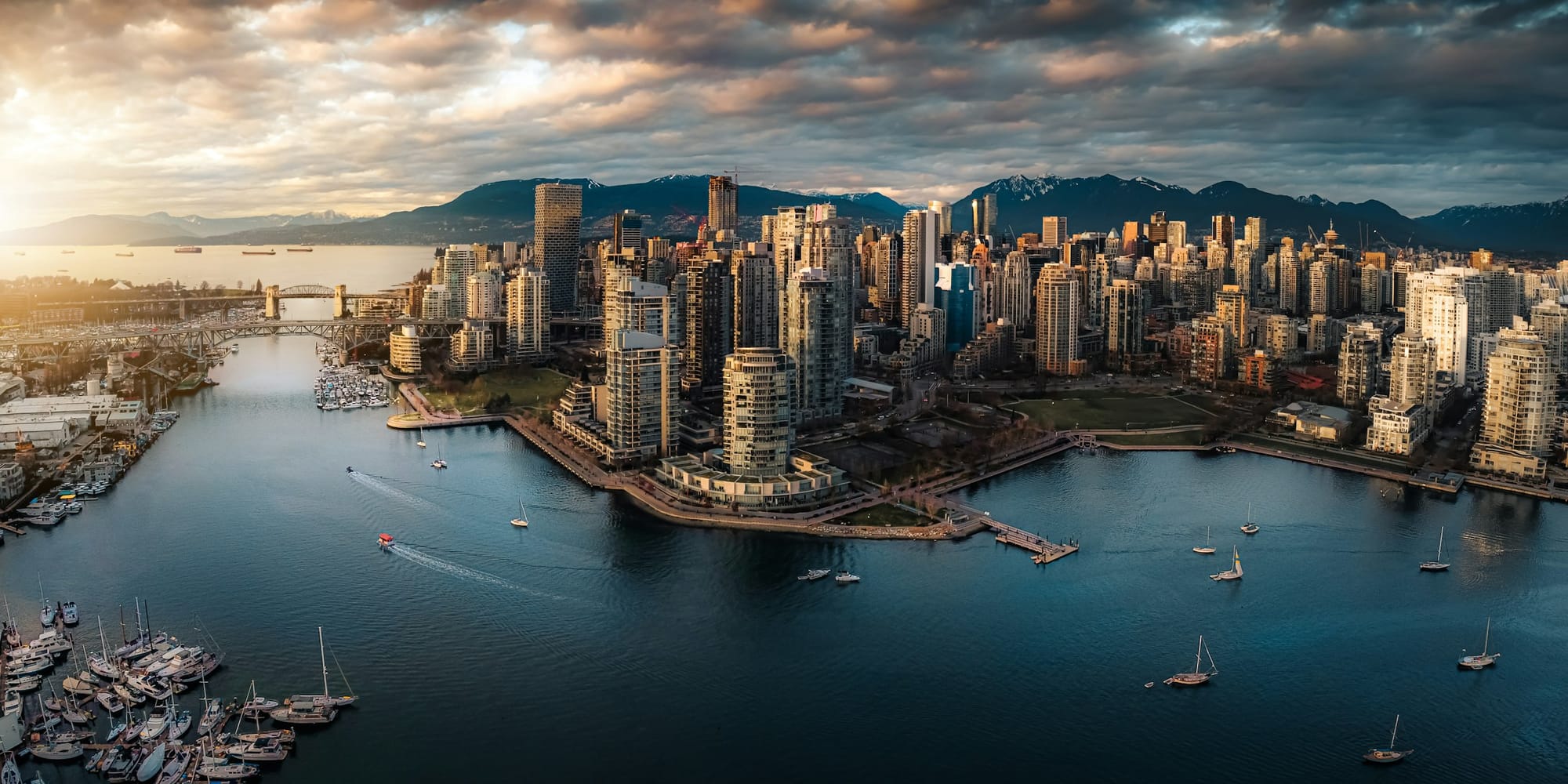 Aerial view of Vancouver — the downtown peninsula, False Creek and the harbour, North Shore mountains rising beyond. The whole hand of the city in one frame.