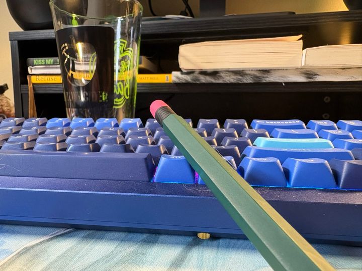 an applce pencil with a green slipcase leaning against a blue mechanical keyboard. A monitor stand is in the background with some books and in front of that is a pint glass.