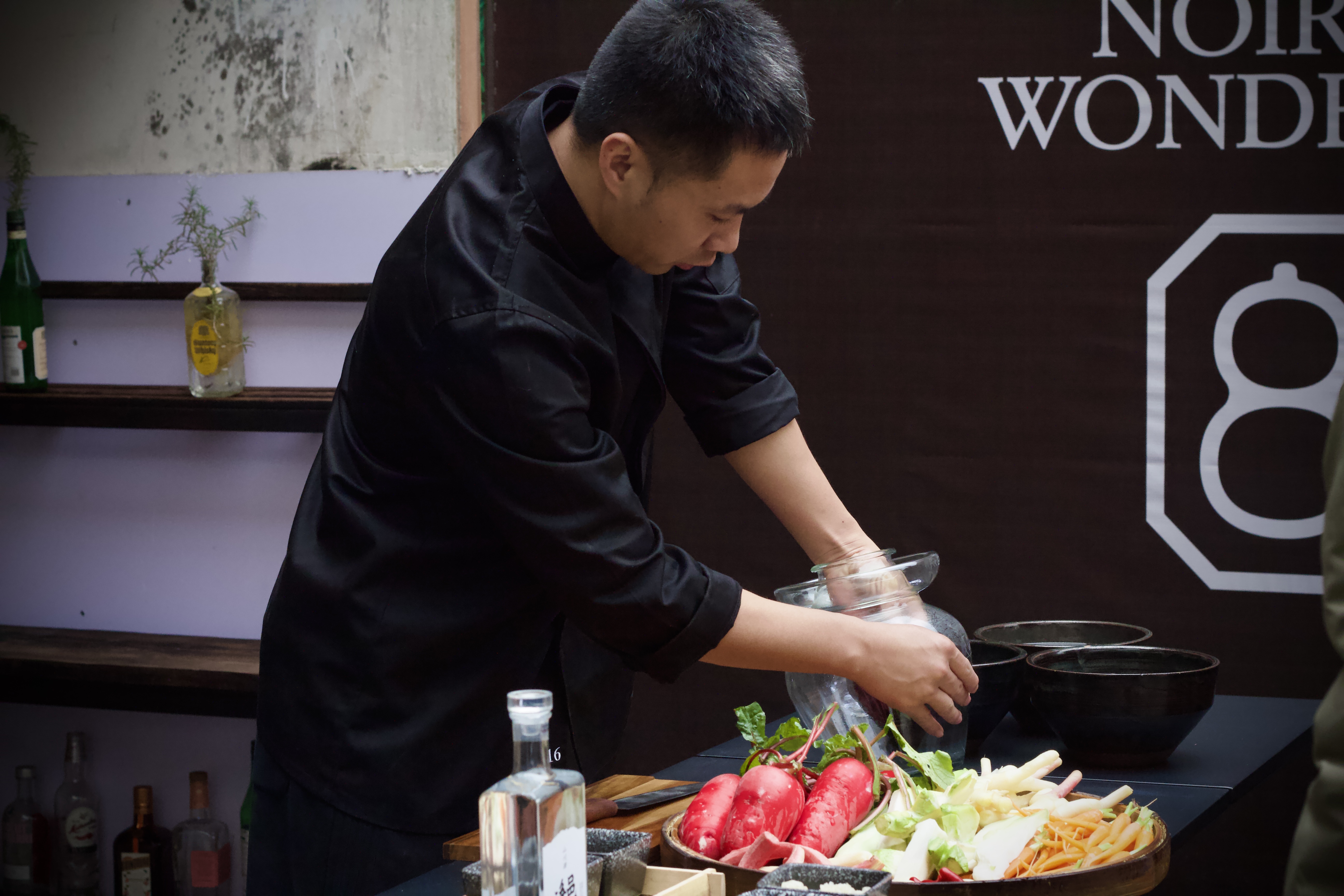 A Chinese Chef (Chef Tang), preparing a pickle jar for a class on teaching pickling, also on the table in front of him are three black bowls, various vegetables, a cutting board and knife, and a bottle of Baijiu.