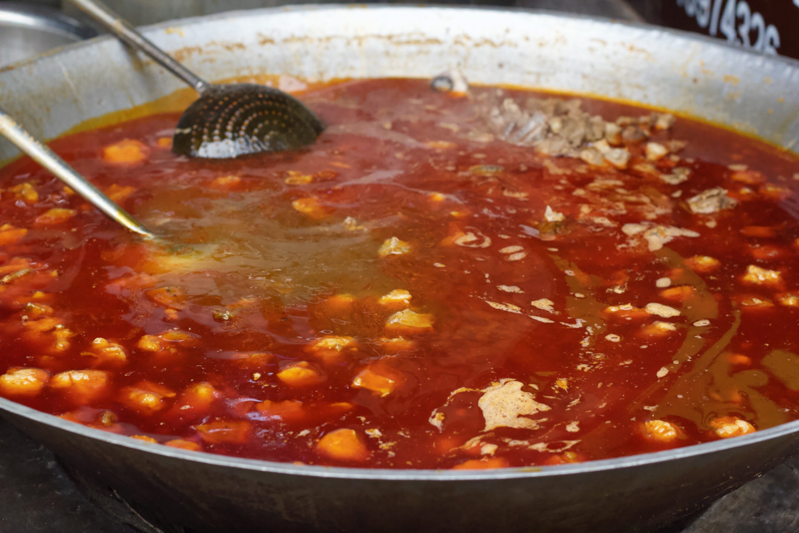 A large steel cooking pot with a deep red broth and floating spices and chunks of lamb meat.