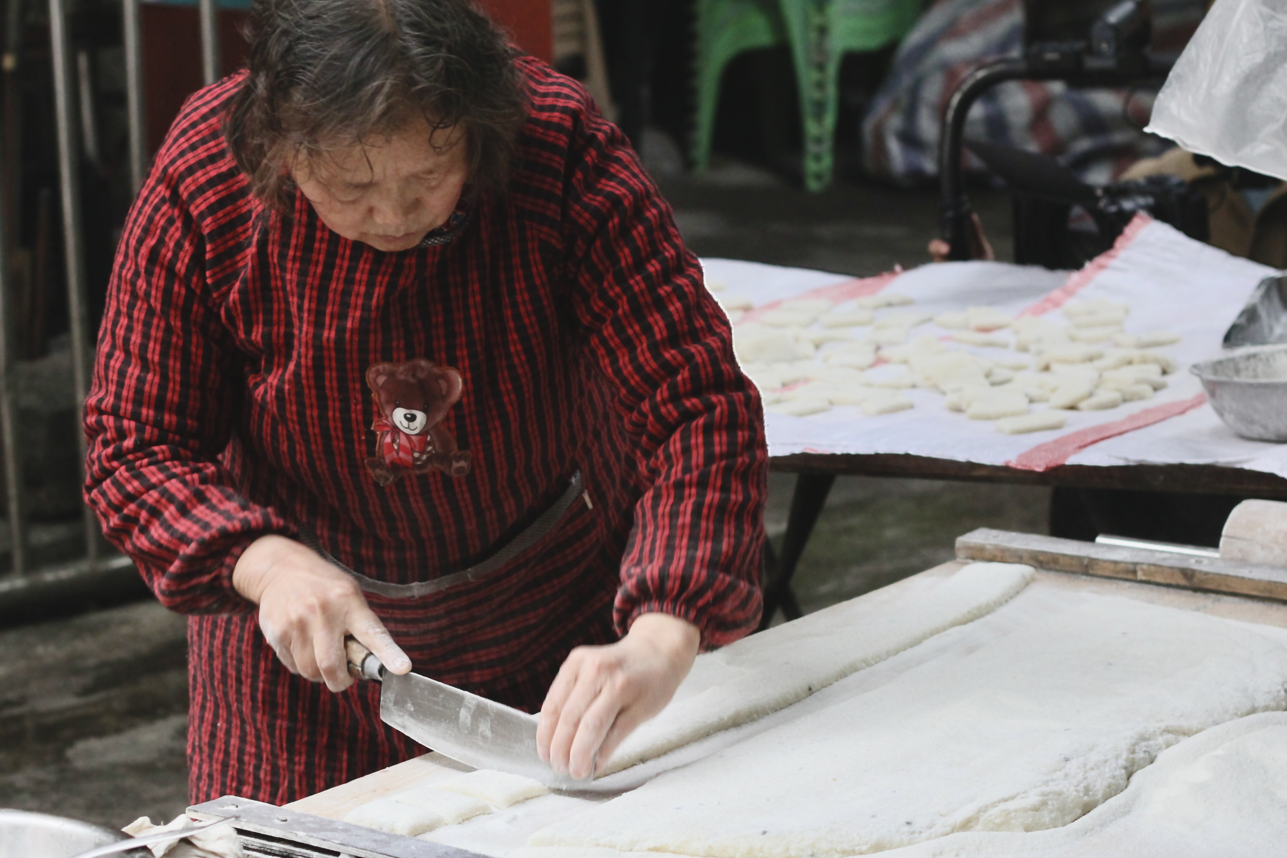 An elderly Chinese woman from the Sichuan province is preparing a traditional rice snack at a street stall in Luzhou. She is using a large knife to cut a long strip into square pieces about 1x2x3cm
