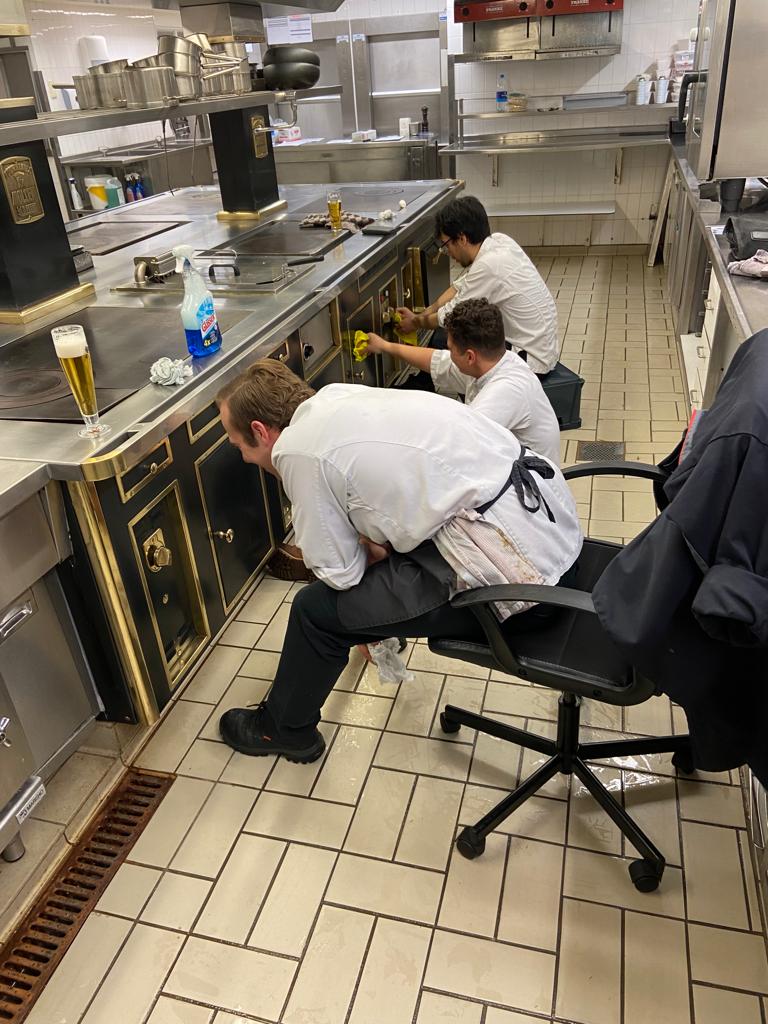 Three chef's cleaning the central cooking hub in a professional restaurant on a beige tiled floor.
